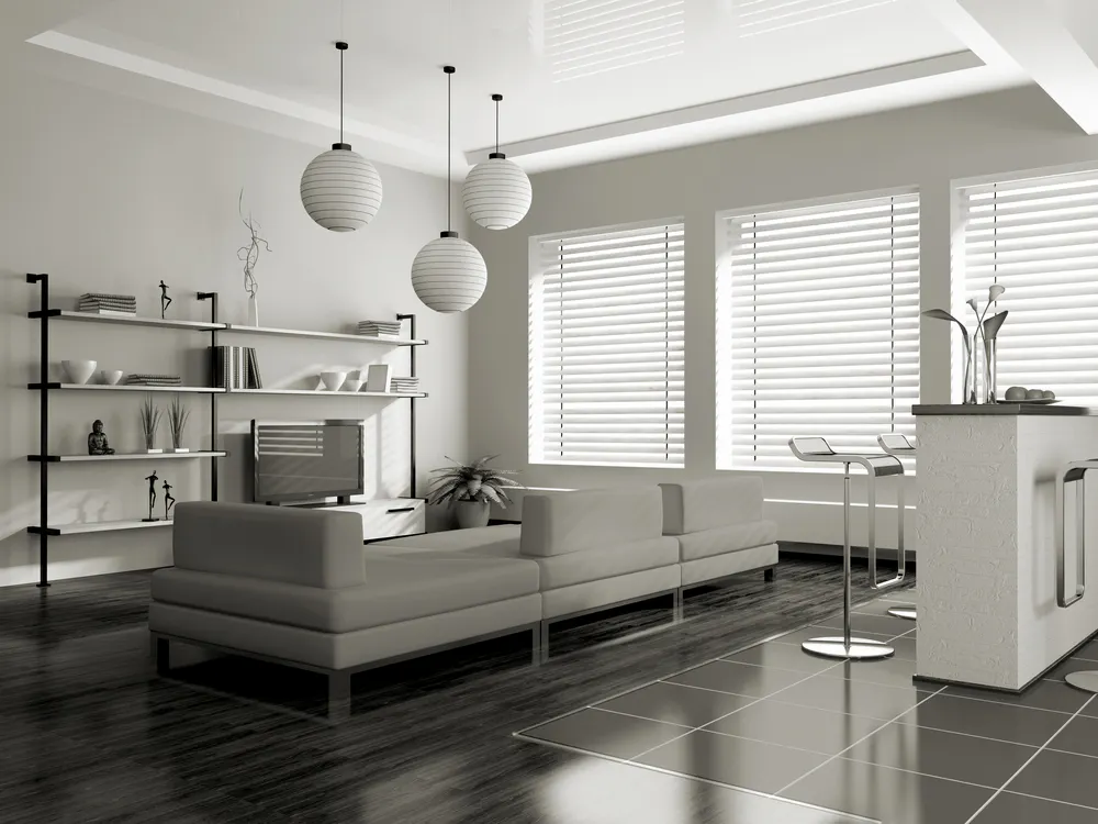 White horizontal window blinds filter sunlight across a modern open-plan living room with low sectional sofa, shelving unit, pendant globe lights and bar stools
