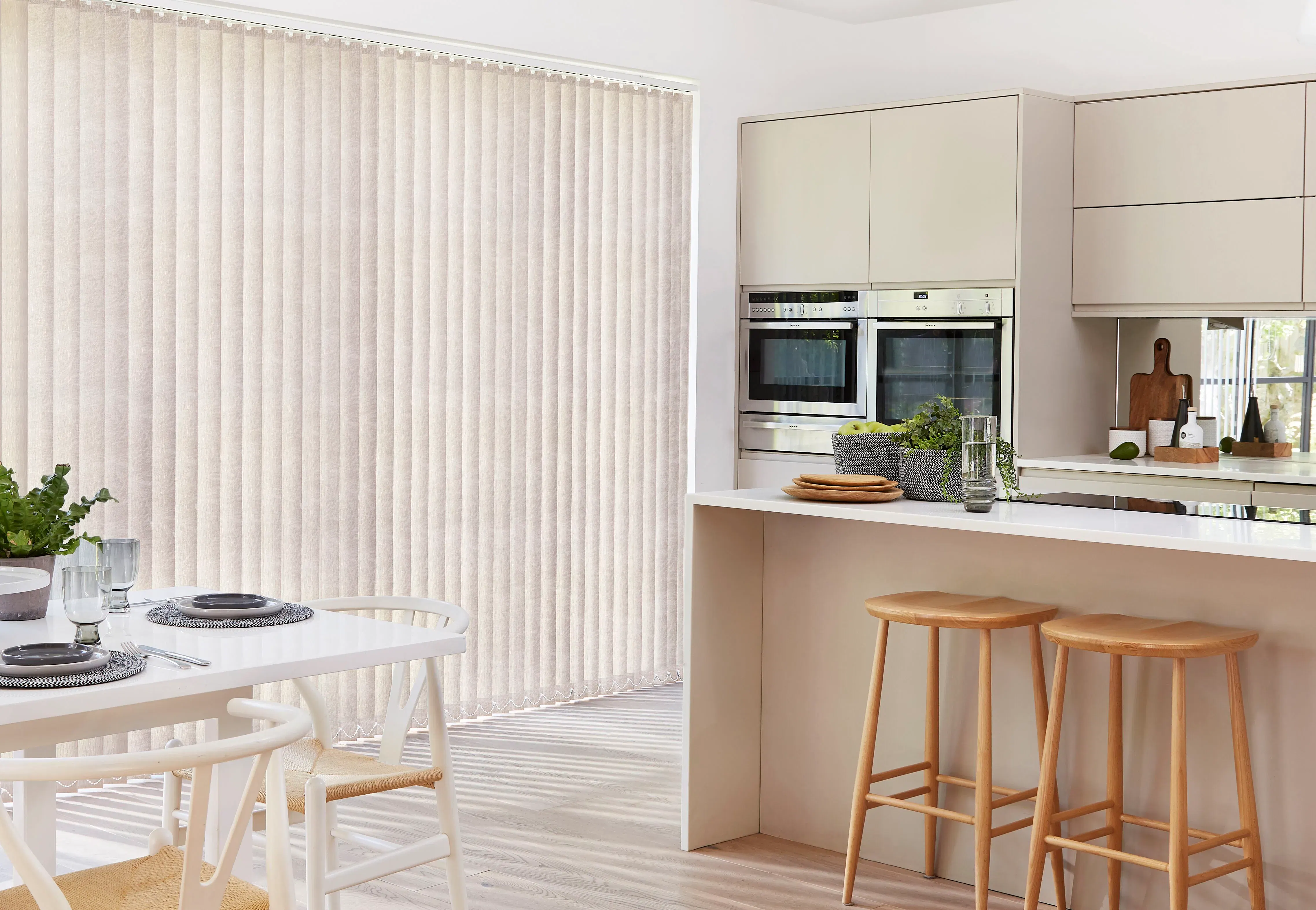 Vertical fabric blinds filtering soft daylight across a modern cream kitchen and dining area with island, wooden bar stools, table place settings and plants