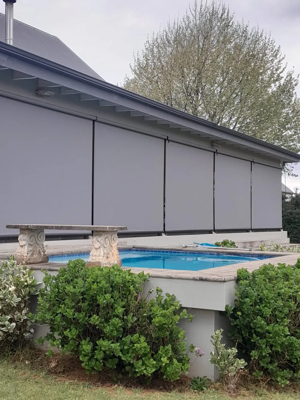 Outdoor roller blinds lowered across a patio, shading a raised swimming pool; stone bench and green shrubs in foreground, house roof and tree behind.