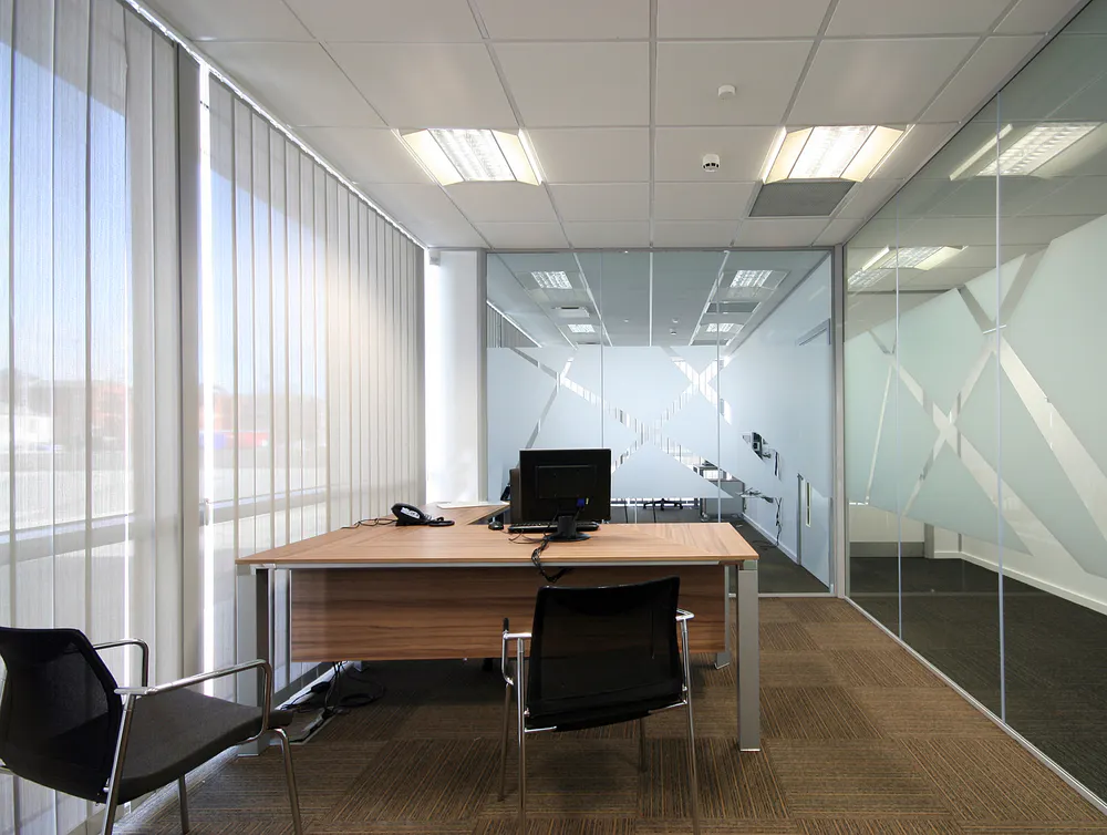 Vertical blinds filtering daylight across a wooden desk with computer and chairs inside a modern glass-walled office, carpeted floor and frosted window graphics