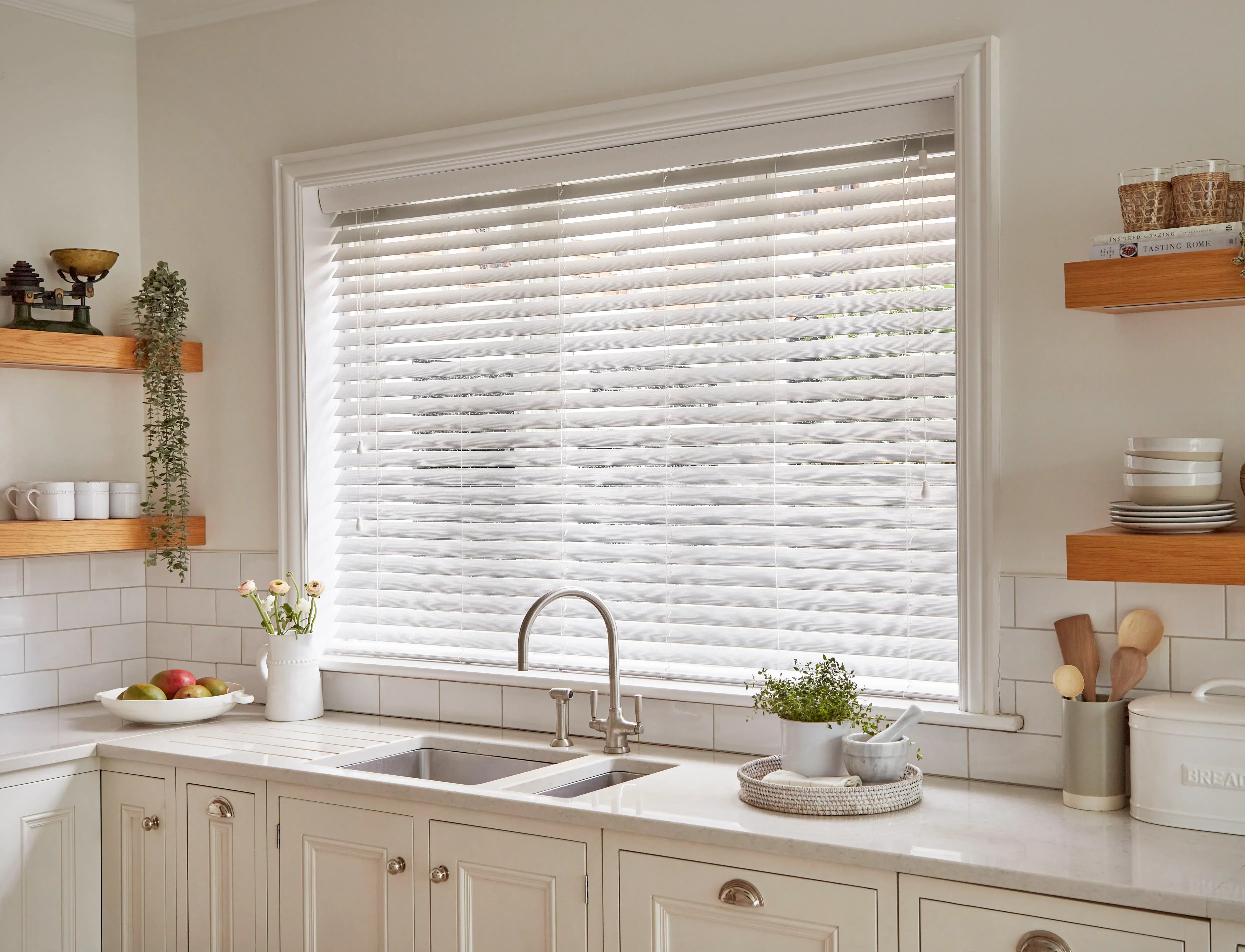 White horizontal window blind tilting closed over a kitchen sink, filtering daylight in a neutral tiled kitchen with wooden shelves, potted plants, mugs and