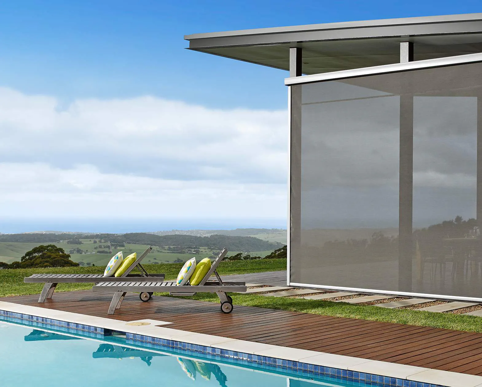 Outdoor roller blind lowered beside a modern patio, shading a glass-walled area by a pool with two loungers and green cushions, overlooking rolling hills and
