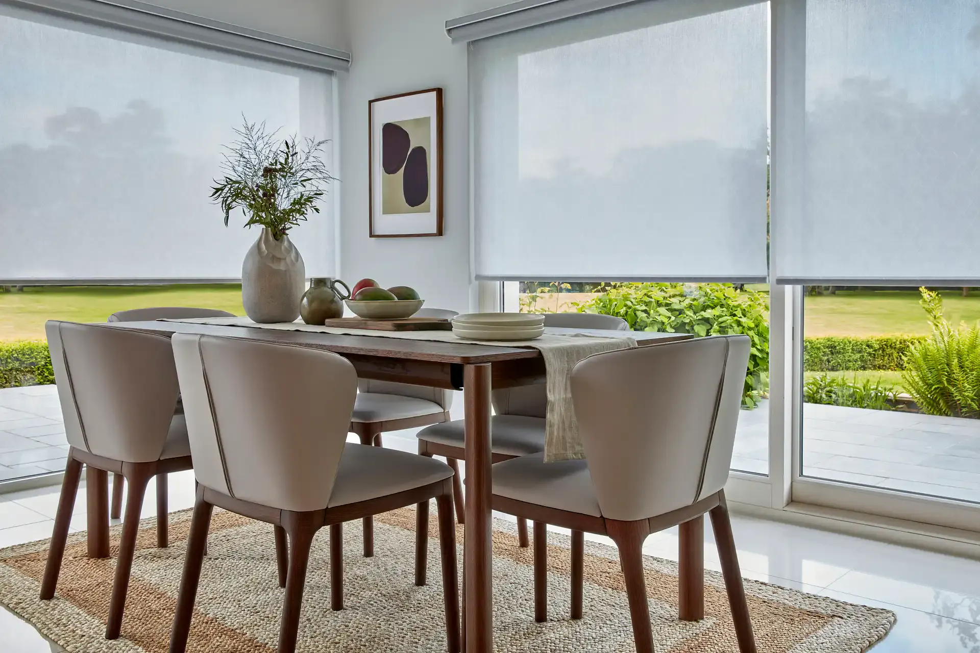 Roller blinds filtering soft daylight across a dining table with chairs, vase and fruit bowl, creating a calm interior view overlooking a neat green garden