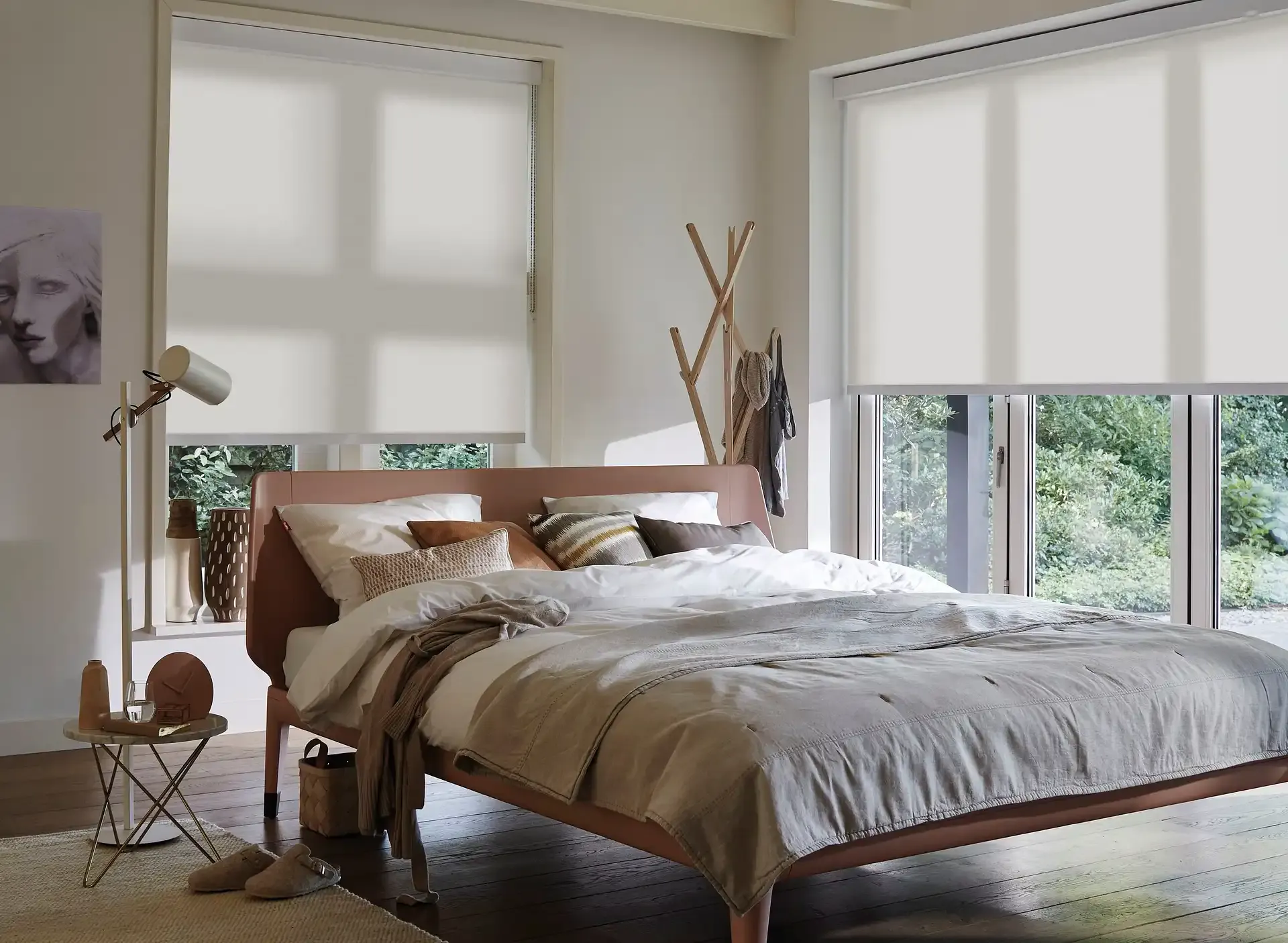 Roller blinds diffuse daylight across a cozy bedroom, covering tall windows above a cushioned bed with pillows and a wooden coat stand, soft greenery visible