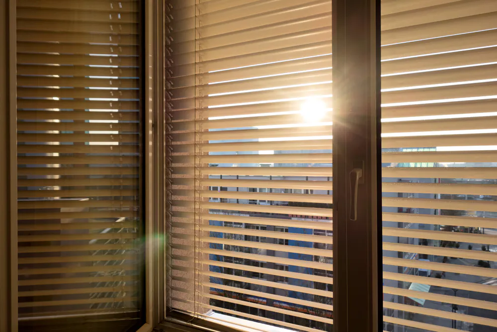 Venetian blinds filtering sunlight across a window, casting horizontal slats' shadows onto the frame and revealing an urban building skyline outside at sunset.