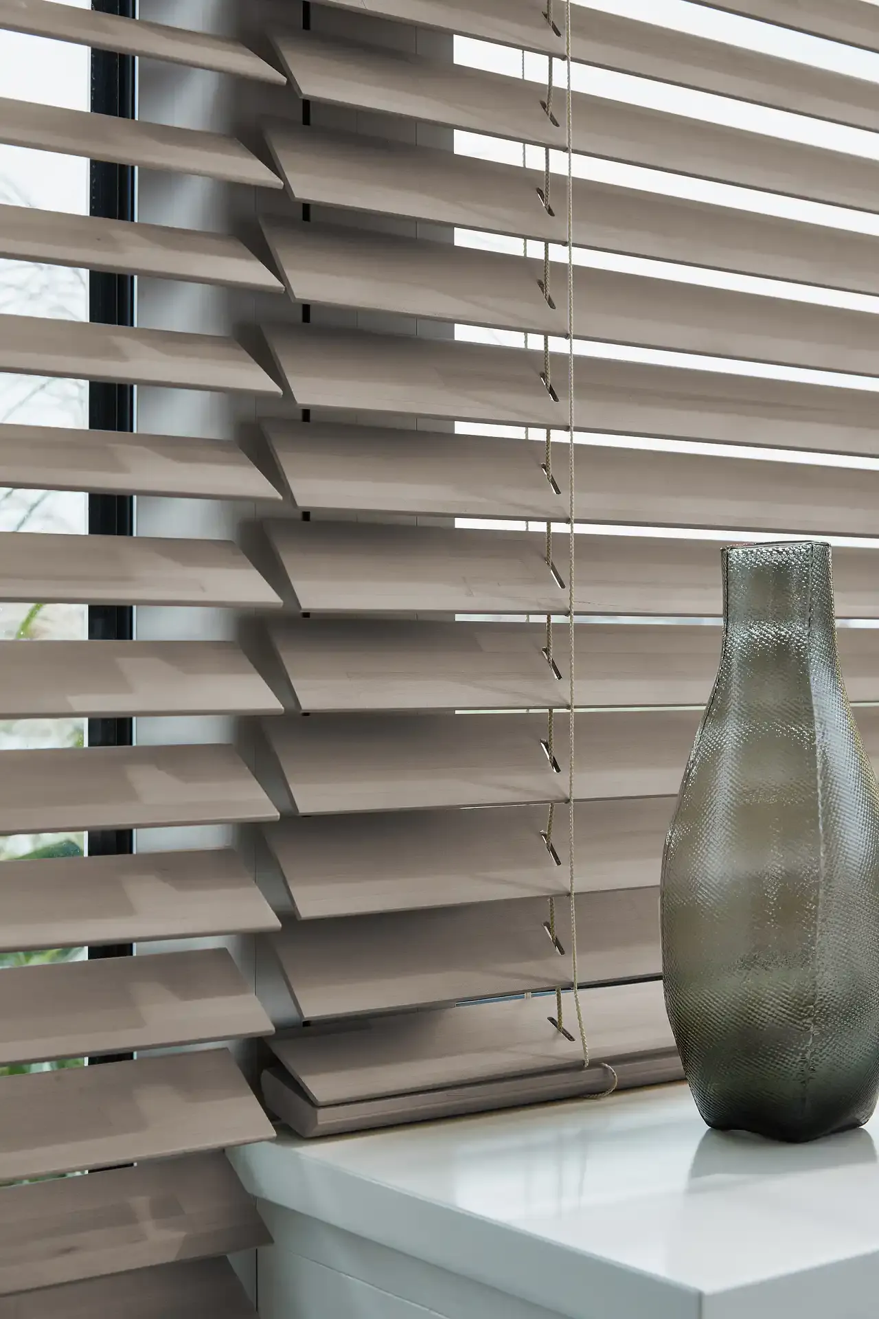 Wooden window blinds with slats tilted open, cords hanging beside a windowsill holding a textured glass vase