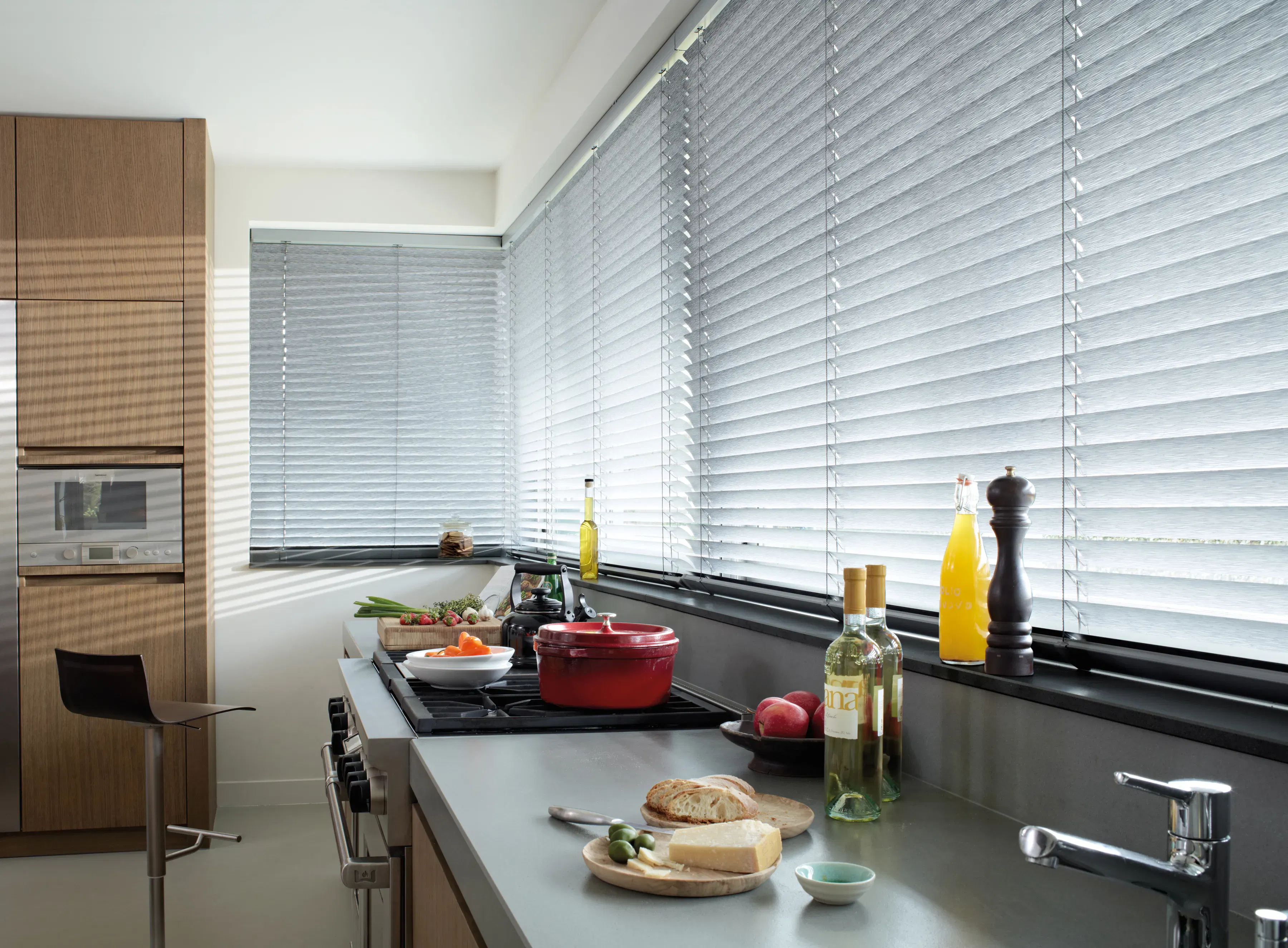 Horizontal silver blinds filter daylight across a modern kitchen, covering corner windows above a countertop with stove, chopping board, bottles and sink