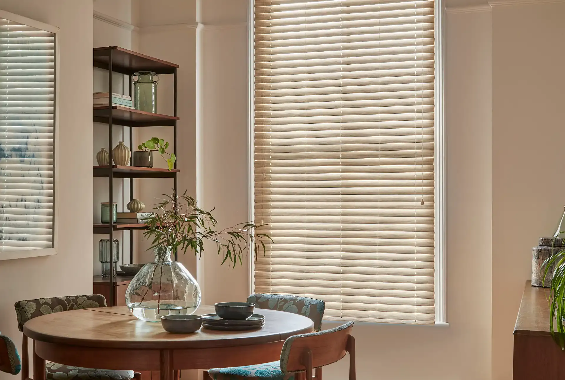 Beige horizontal window blind filters soft daylight into a cozy dining room, beside a round wooden table with bowls, glass vase, shelving with decorative vases