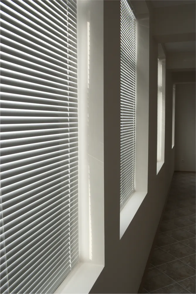 Venetian blinds filtering sunlight across recessed windows in a narrow tiled corridor, casting parallel light-and-shadow lines along white walls and sills.
