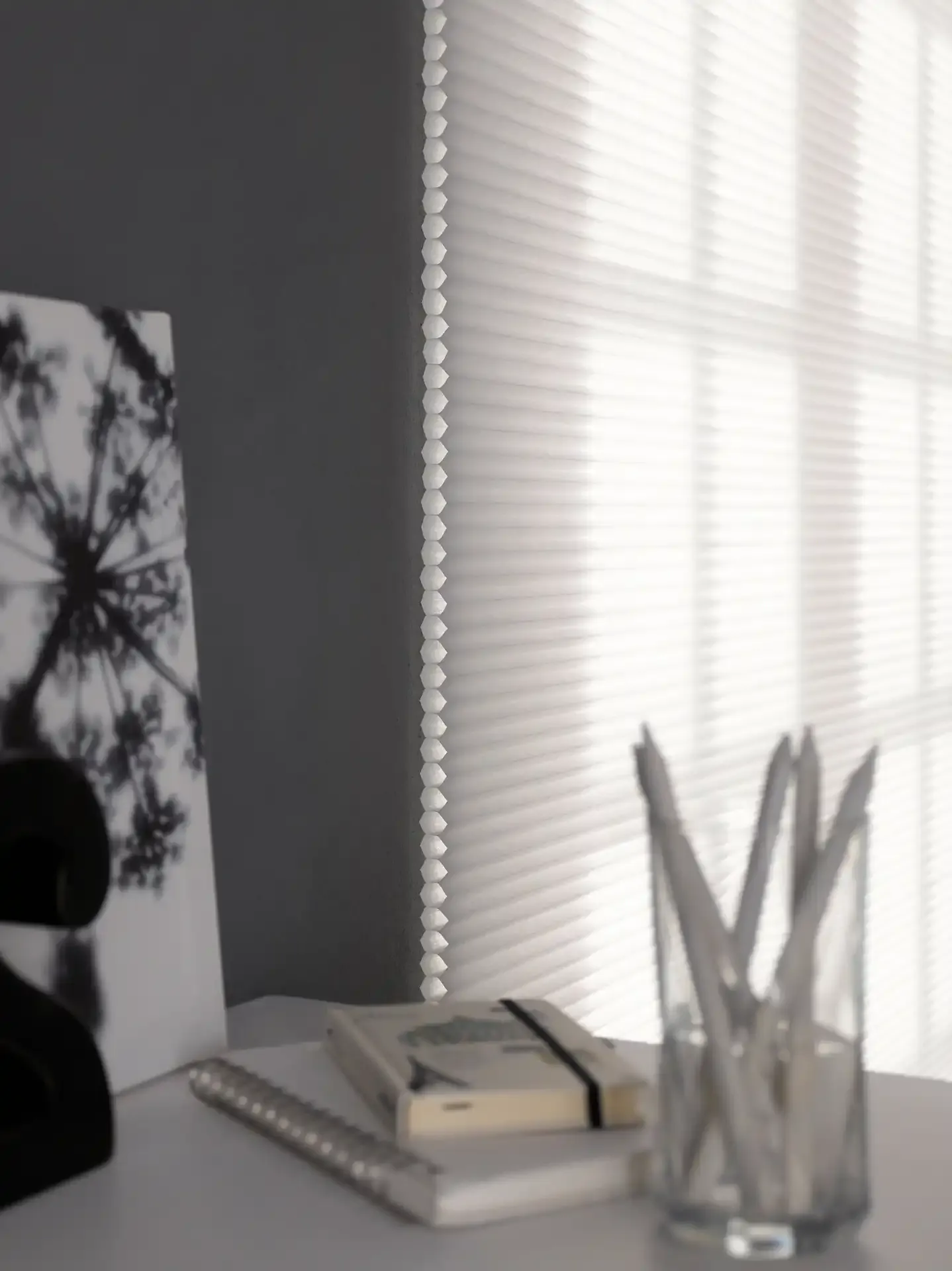 Honeycomb blind filters soft daylight across a desk, casting striped shadows; notebooks and a glass pen holder sit nearby in a minimalist indoor workspace