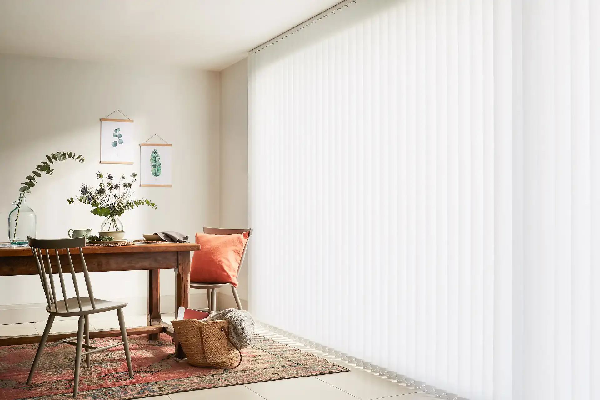 Vertical blinds filter soft daylight across a cozy dining nook with wooden table, vase of foliage, chairs, orange cushion and a woven basket on a patterned rug