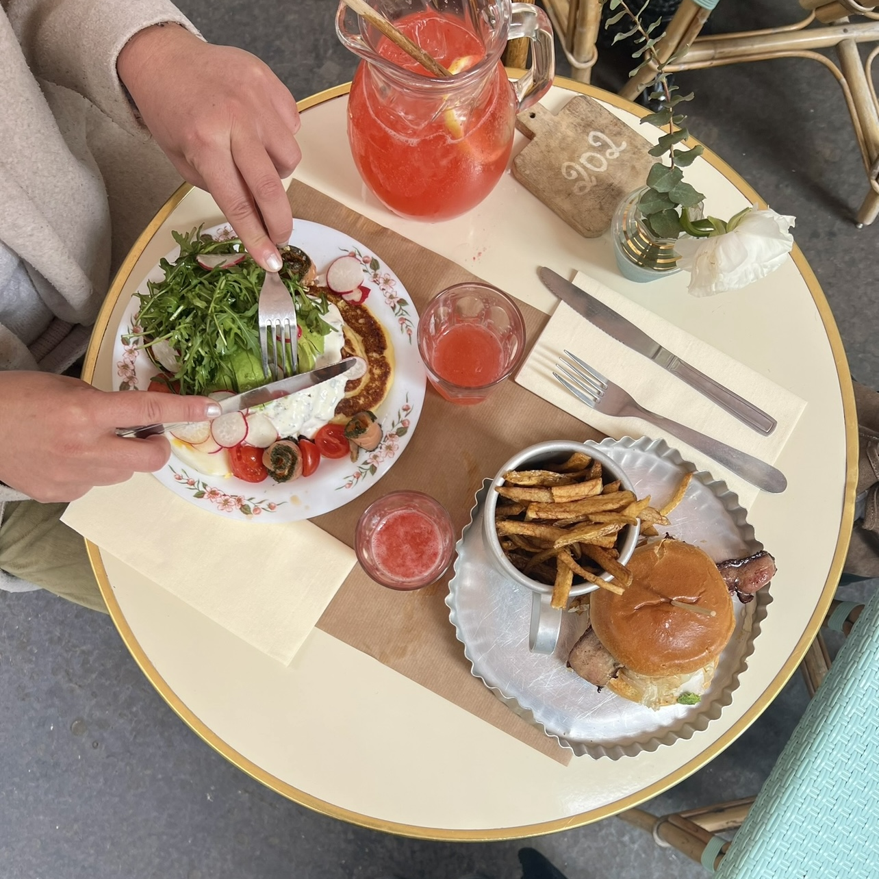 Table beige avec deux plats, un burger frites et des pancakes au saumon et avocat. Avec deux verres de limonades rouge. Et une personne coupant ses pancakes.