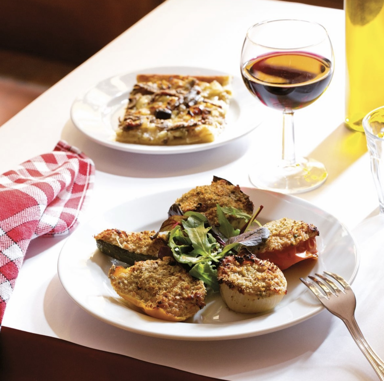 Table comprenant deux assiettes, l'une avec une part de pissaladière à l'intérieur. L'autre avec des petits farcies. Ainsi qu'un verre de vin