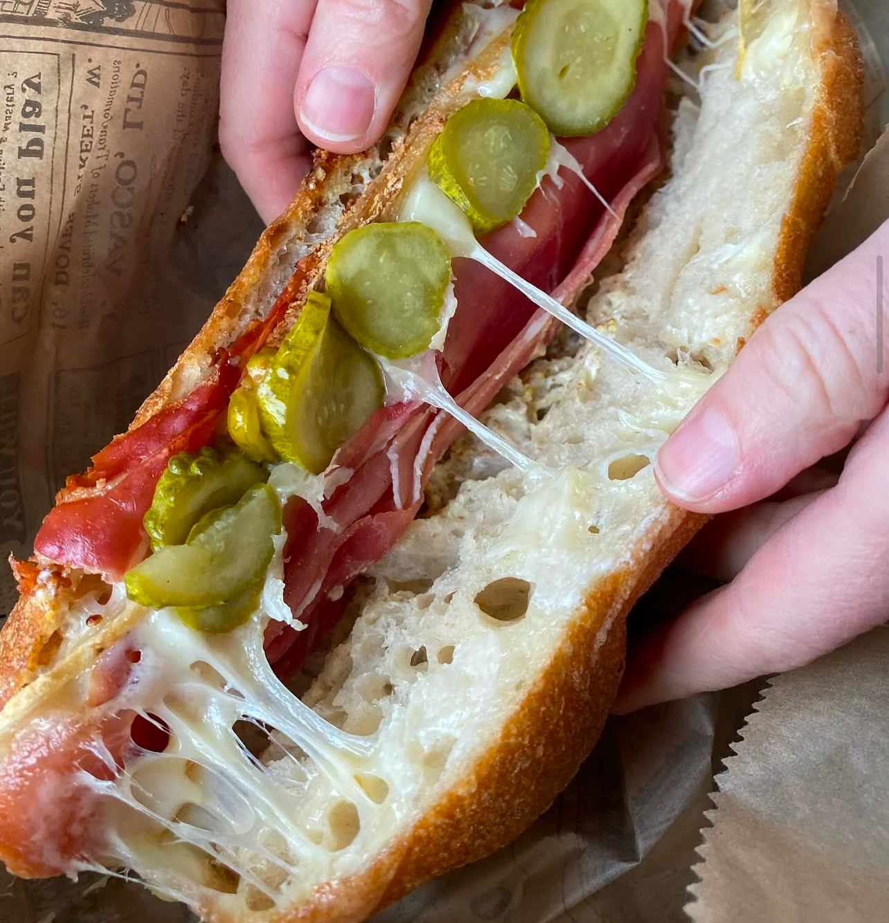 Mains ouvrant un sandwich au jambon, cornichons et fromage fondu
