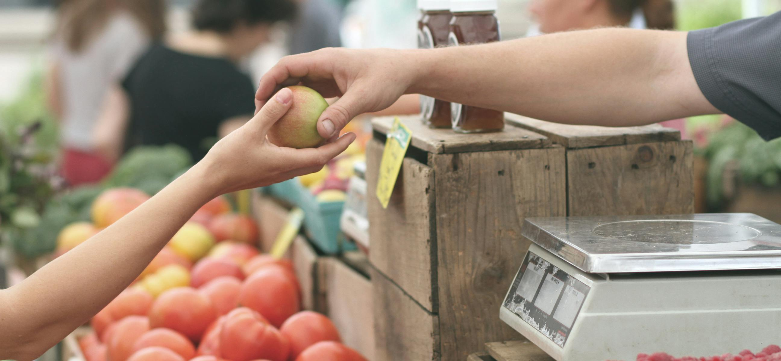 Une main donne une pomme à une autre main au dessus d'une étale de fruits.
