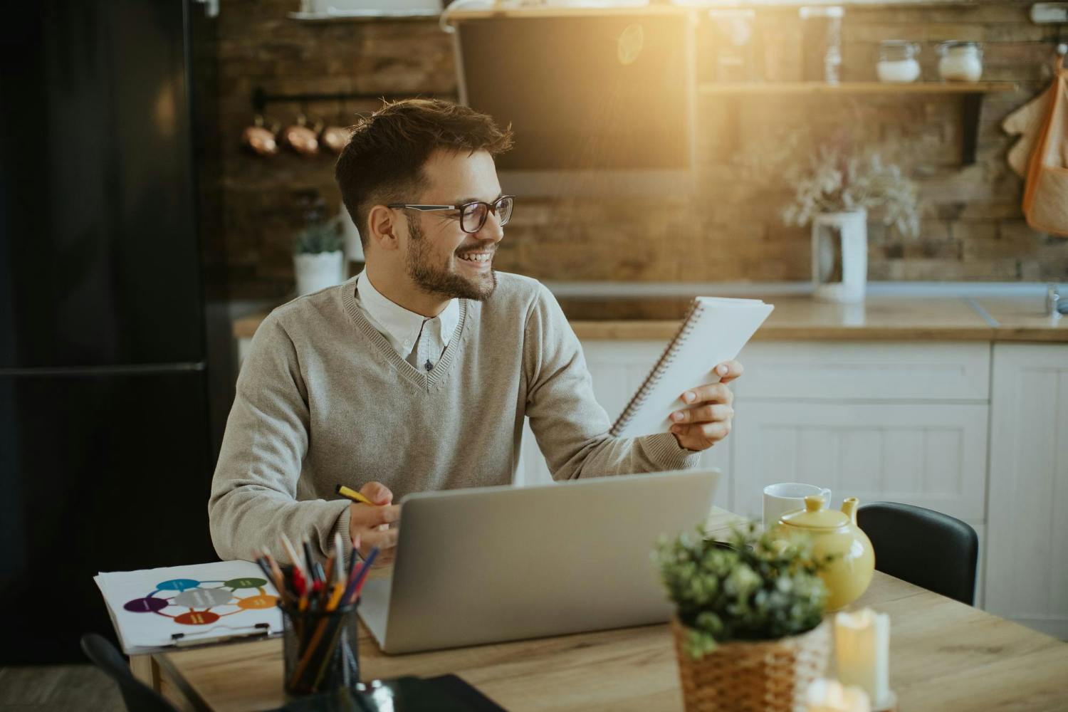 Homem sorrindo, em uma cozinha, sentado à mesa com um notebook a sua frente e fazendo anotações em um pequeno caderno .