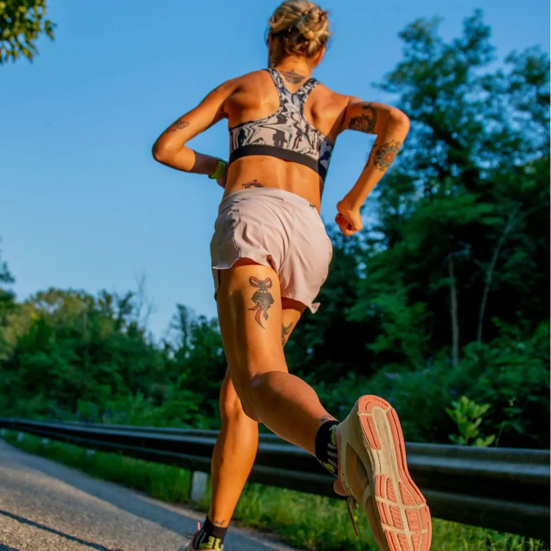 Mulher praticando corrida ao ar livre em trilha na natureza, vestida com roupas esportivas, sob céu azul claro e árvores verdes ao fundo, promovendo saúde e bem-estar.