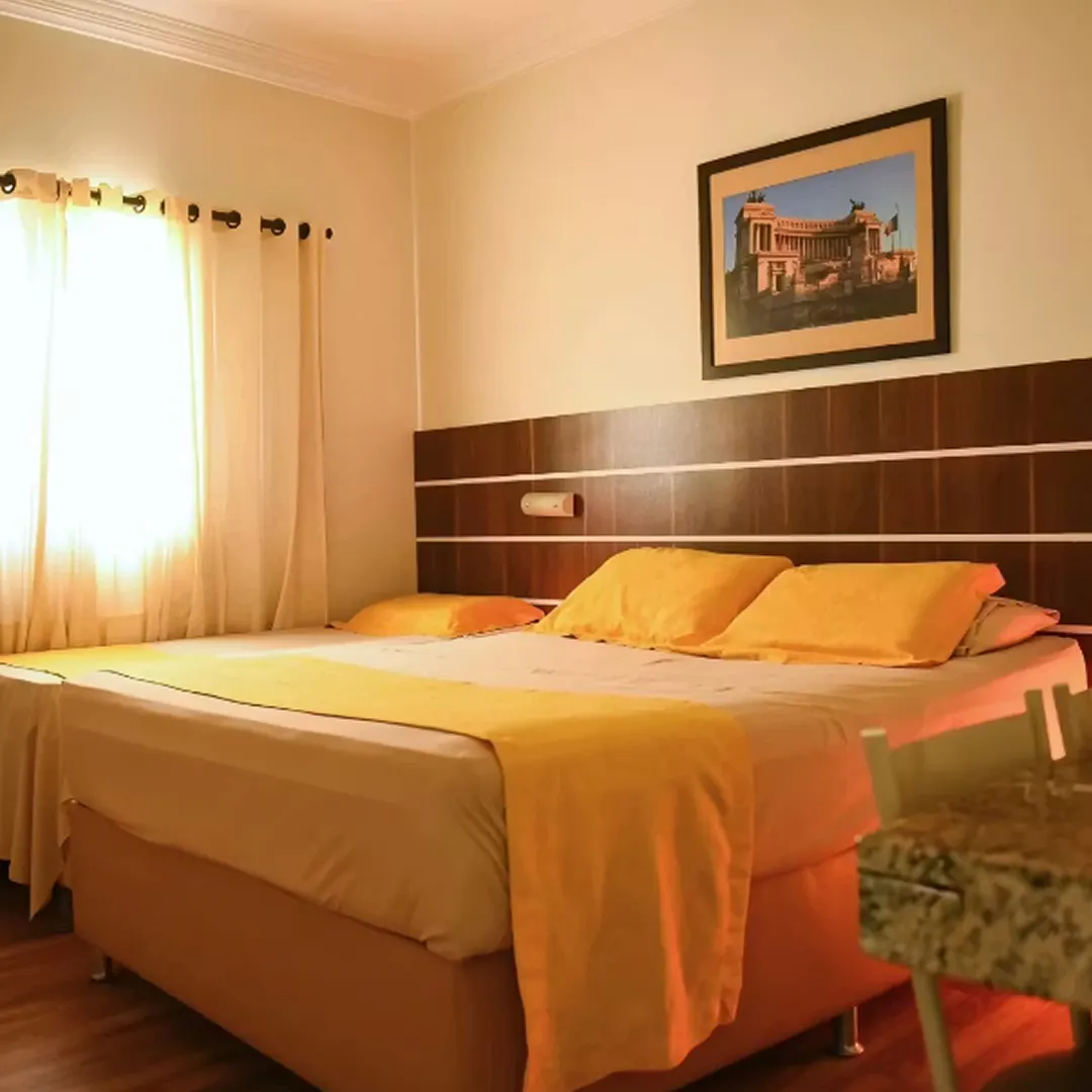 Quarto de hotel confortável com cama de casal, roupas de cama amarelas, janela com cortinas de voal e decoração moderna na parede.