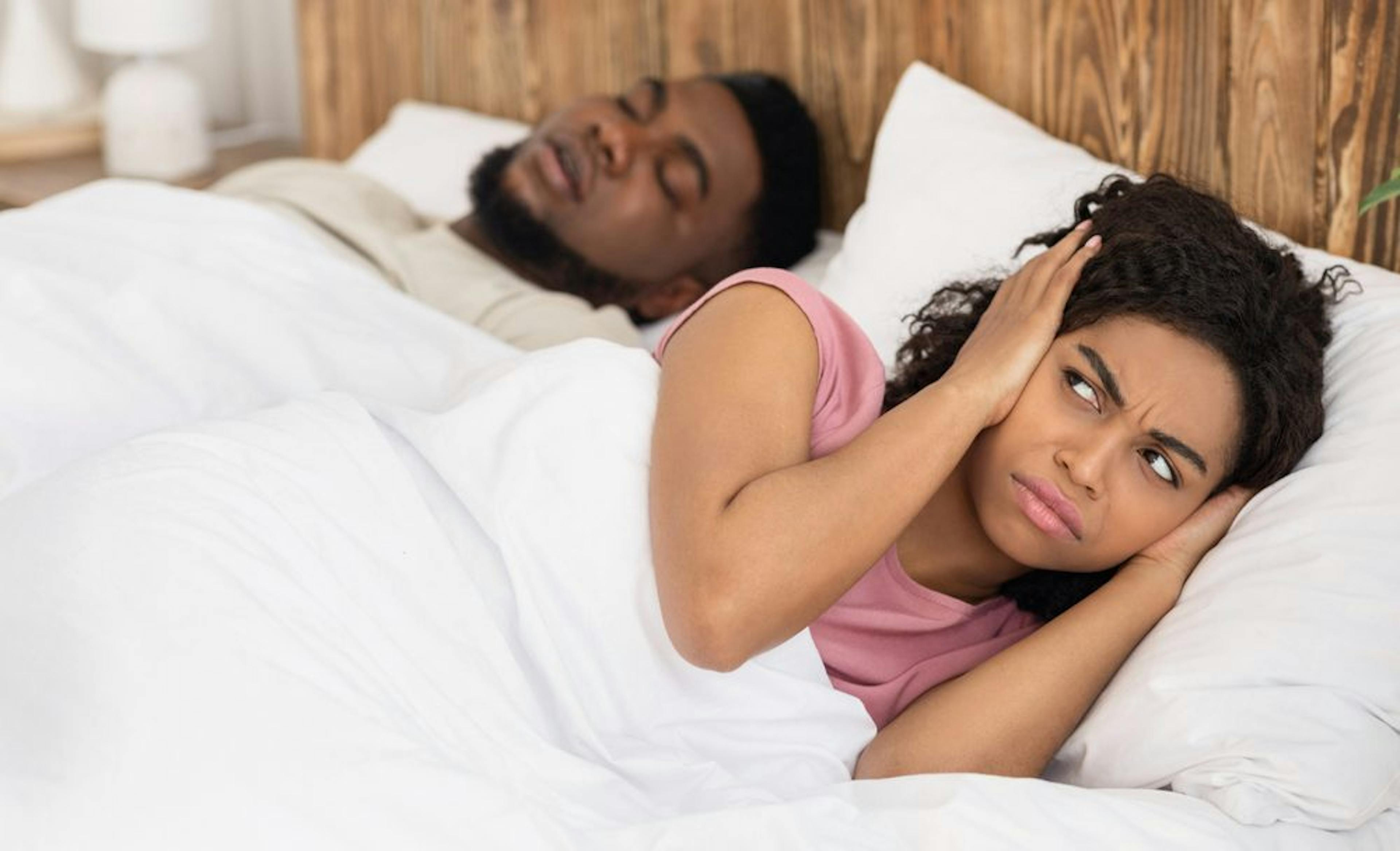 Um casal de pessoas negras deitados na cama. O marido, dormindo e roncando, e sua esposa acordada com as mãos em seus ouvidos.