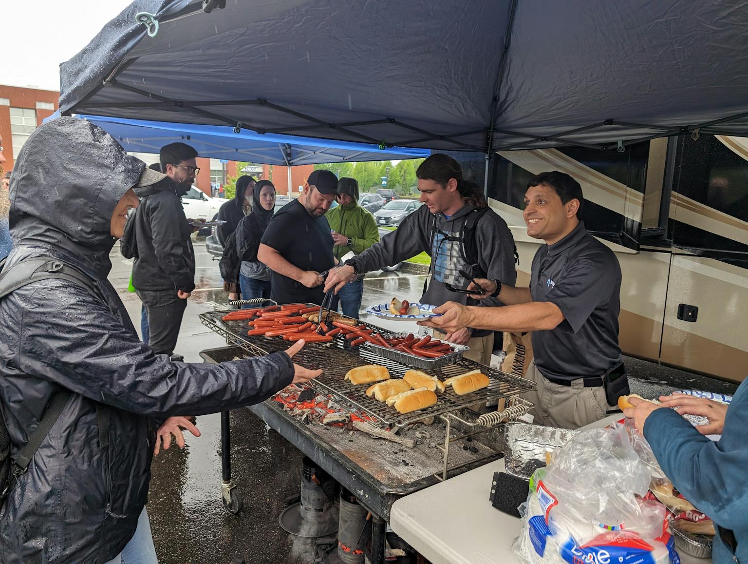 Noah smiling while serving hot dogs with Jeff and Alex also manning the grill in front of Lady Jupiter. In the rain.