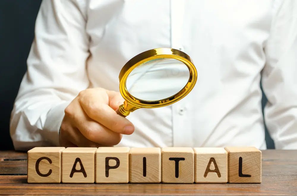 Man with a magnifying glass pointed at the word "Capital"