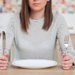 Foto de mulher sentada em uma mesa com um prato de comida vazio segurando o garfo e faca.