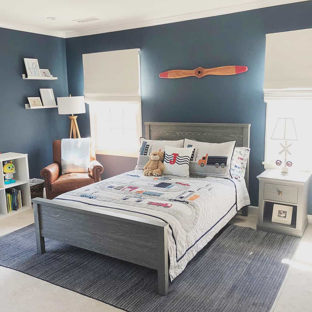 Planes, Trains and automobiles themed boy's room, dark blue walls and off white roman shades on either side of a grey bedframe.
