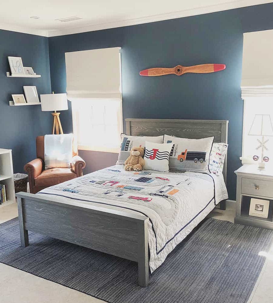 Planes, Trains and automobiles themed boy's room, dark blue walls and off white roman shades on either side of a grey bedframe.