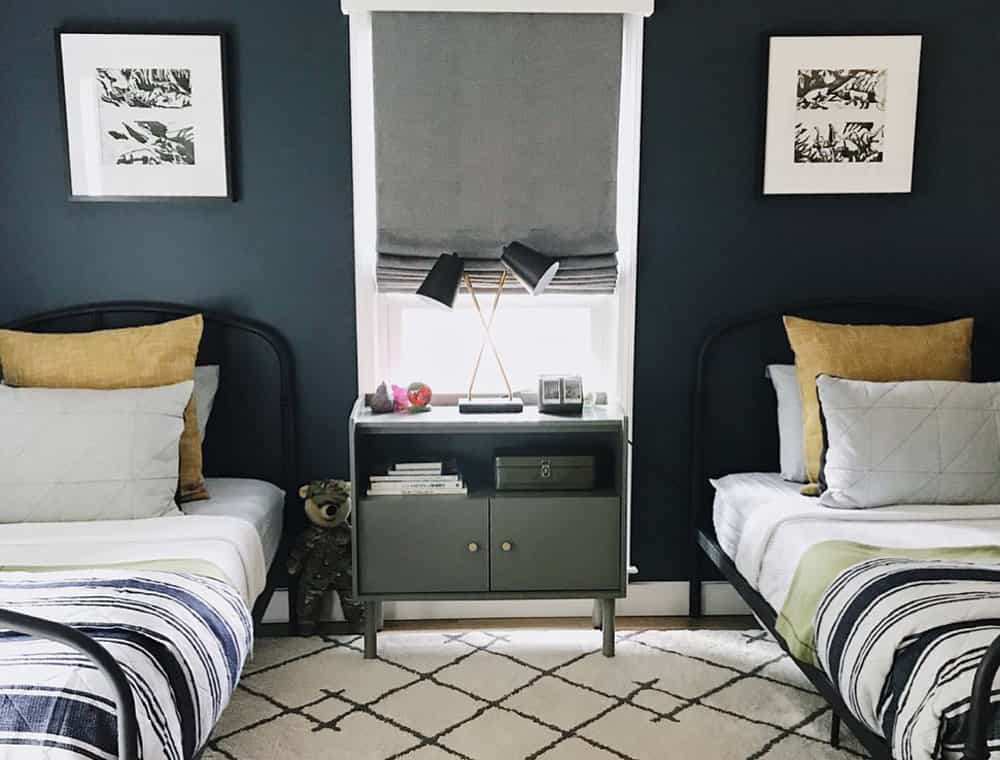 Boys bedroom with two twin beds against a navy blue wall and a grey roman shade in the window in the middle.