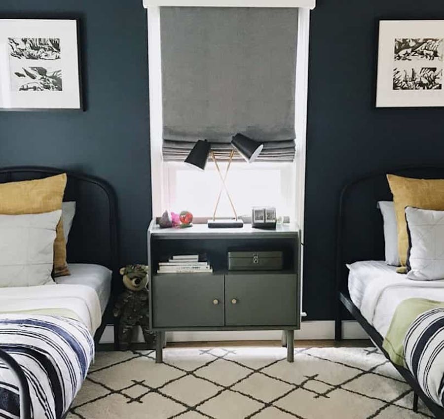 Boys bedroom with two twin beds against a navy blue wall and a grey roman shade in the window in the middle.