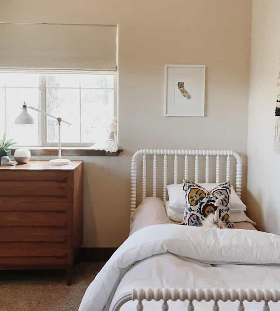 Girl's room with neutral pink and white bedding, wall tapestry, and off white roman shades in the window.