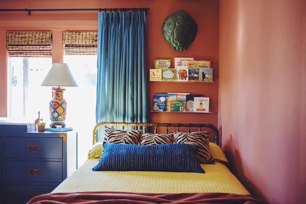 Yellow bed pushed into a corner with terra cotta colored walls, contrasting blue drapes and woven wood shades on the window. Children's books mounted on a shelf above the bed.