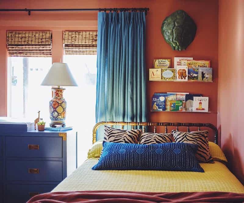 Yellow bed pushed into a corner with terra cotta colored walls, contrasting blue drapes and woven wood shades on the window. Children's books mounted on a shelf above the bed.
