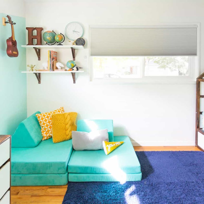 Kids bedroom corner with modular turquoise sofa, yellow pillows, shelf with different sized globes and a grey cellular shade in the window.