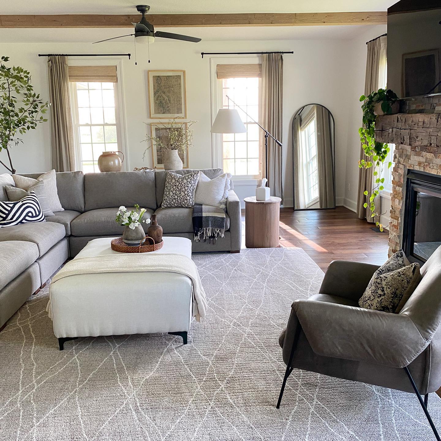 A fall home with a modern living room with faux plants and a floral centerpiece on the ottoman. There are woven wood shades from Blinds.com covering the windows.