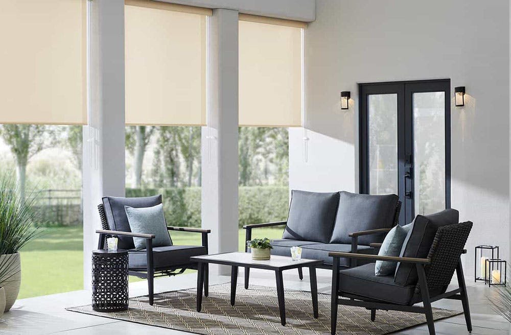 outdoor patio with grey, cushioned chairs around a coffee table and beige solar shades shielding the chairs from direct sun.