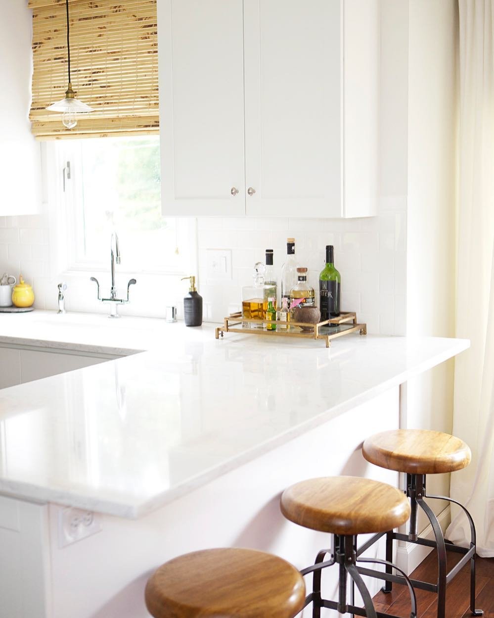 Kitchen area with bright, white countertops and wooden stools. Woven Wood Shades are on the window above the sink.