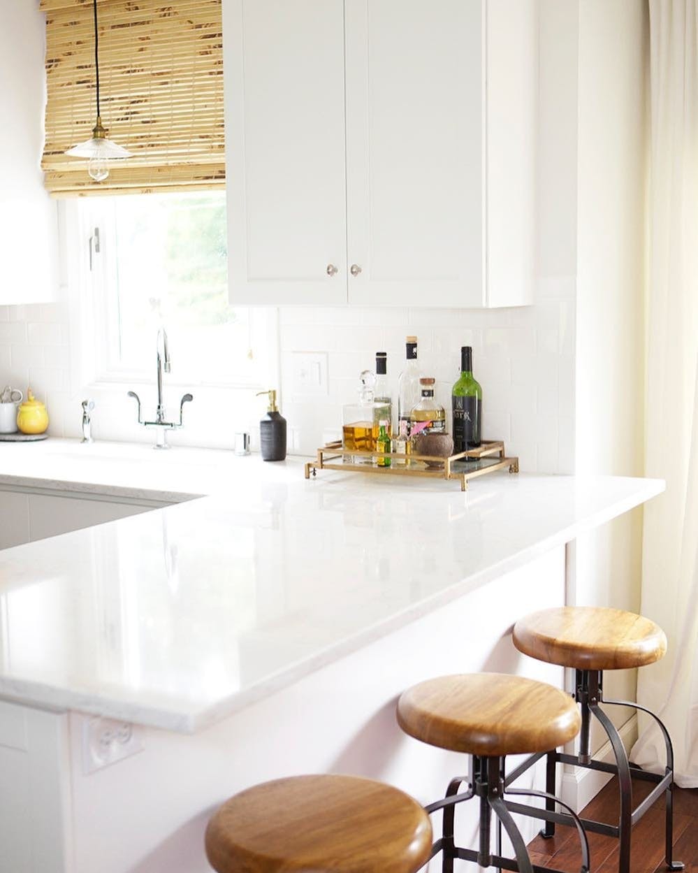 Kitchen area with bright, white countertops and wooden stools. Woven Wood Shades are on the window above the sink.