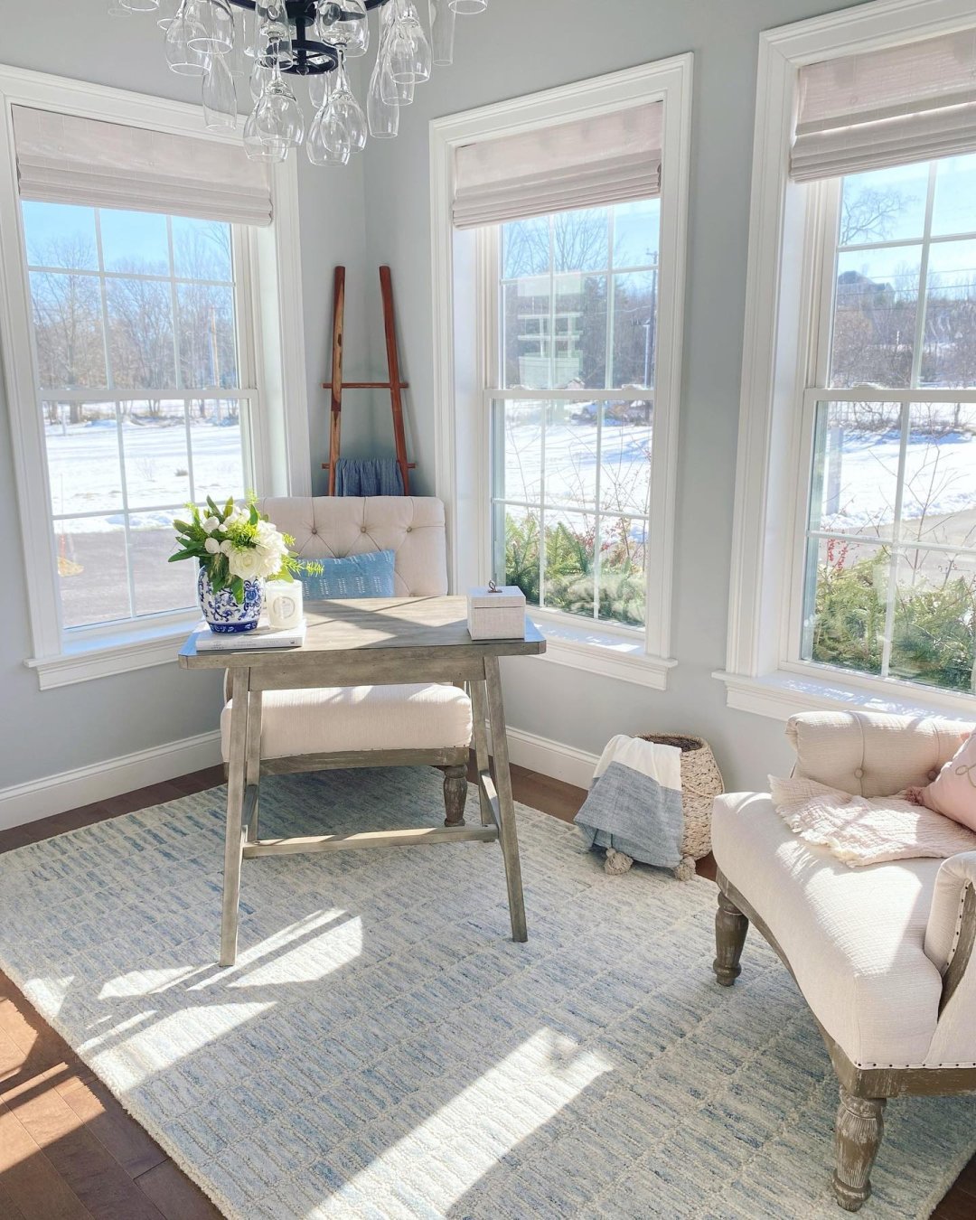 Sitting area with beige chairs and a grey, wooden desk. The walls are painted pale blue-grey and white Woven Wood Shades are on the windows.