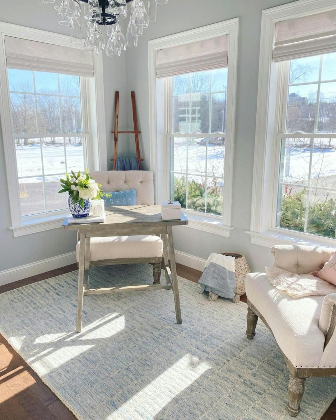 Sitting area with beige chairs and a grey, wooden desk. The walls are painted pale blue-grey and white Woven Wood Shades are on the windows.