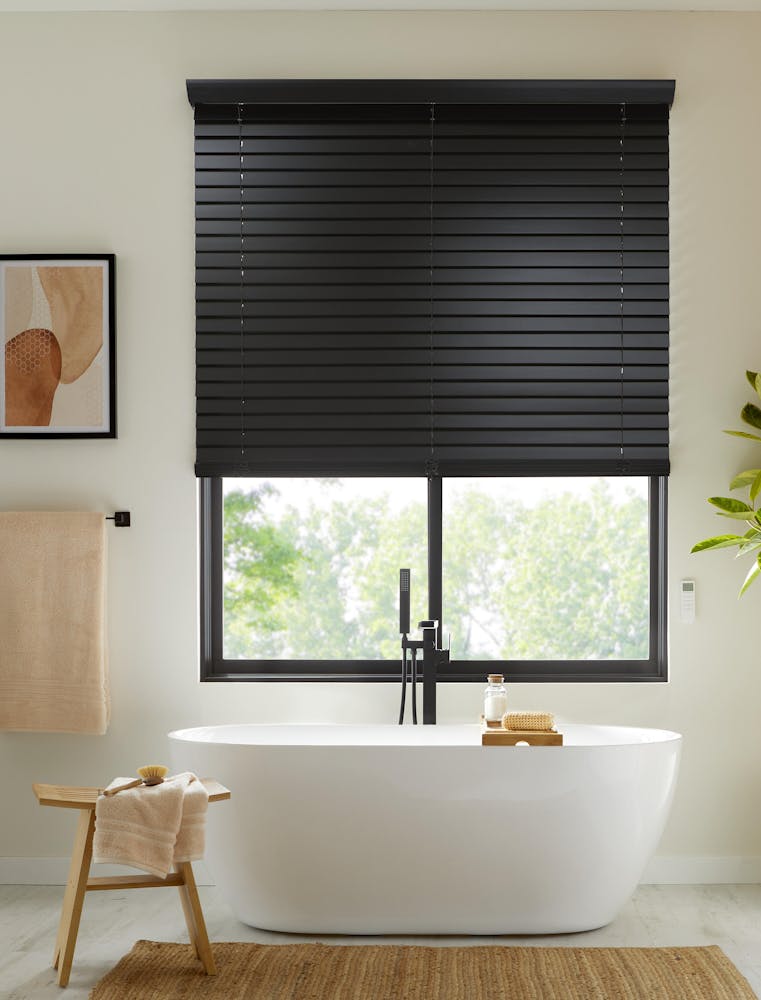 Modern bathroom with Blinds.com Premium 2.5 inch Wood Blinds in black. There is a towel rack with a peach towel next to the  bathtub and small stool with a peach towel draped over it. 