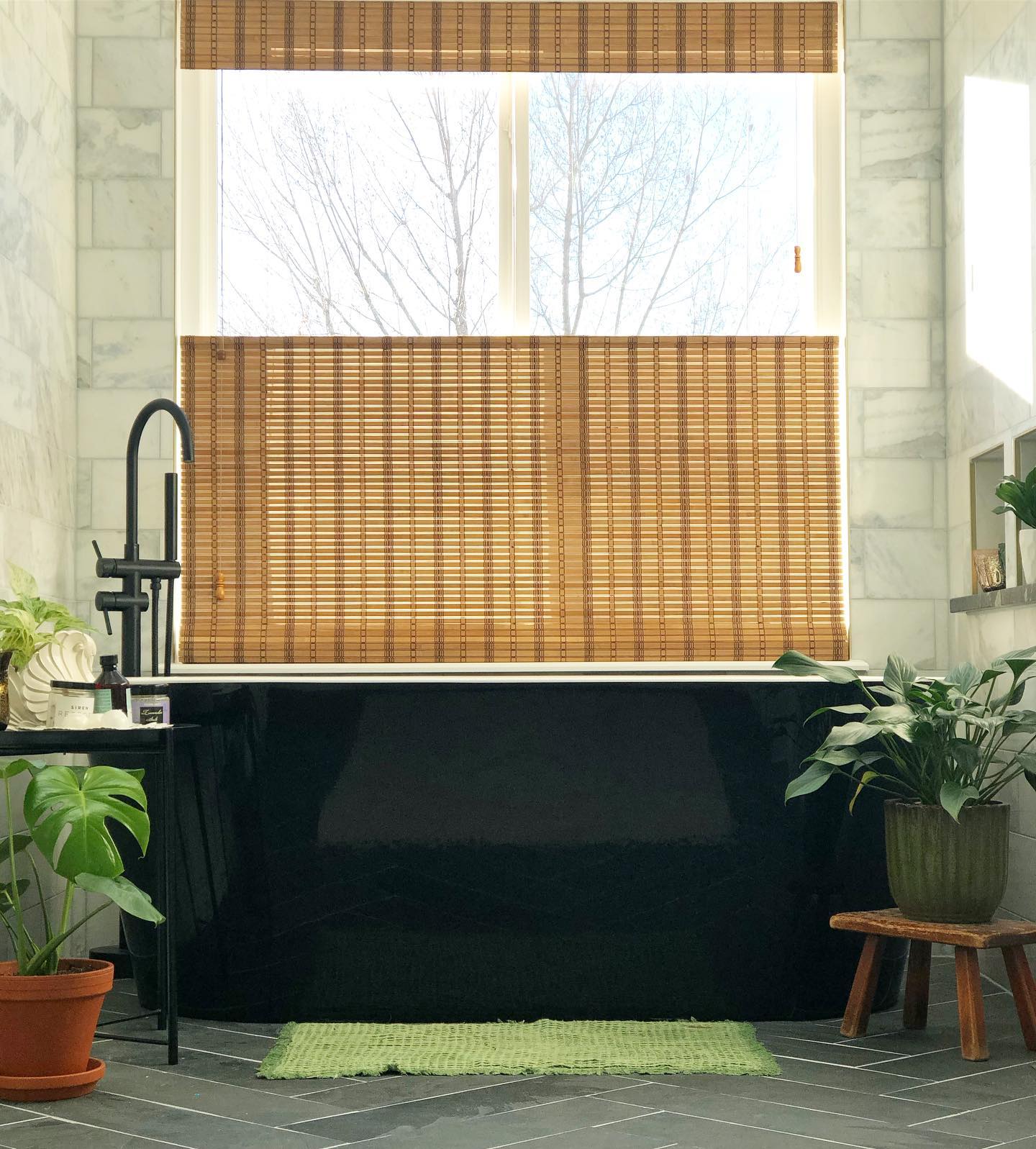 Large, black, free standing tub next to a window with a woven wood shades and surrounded by plants.