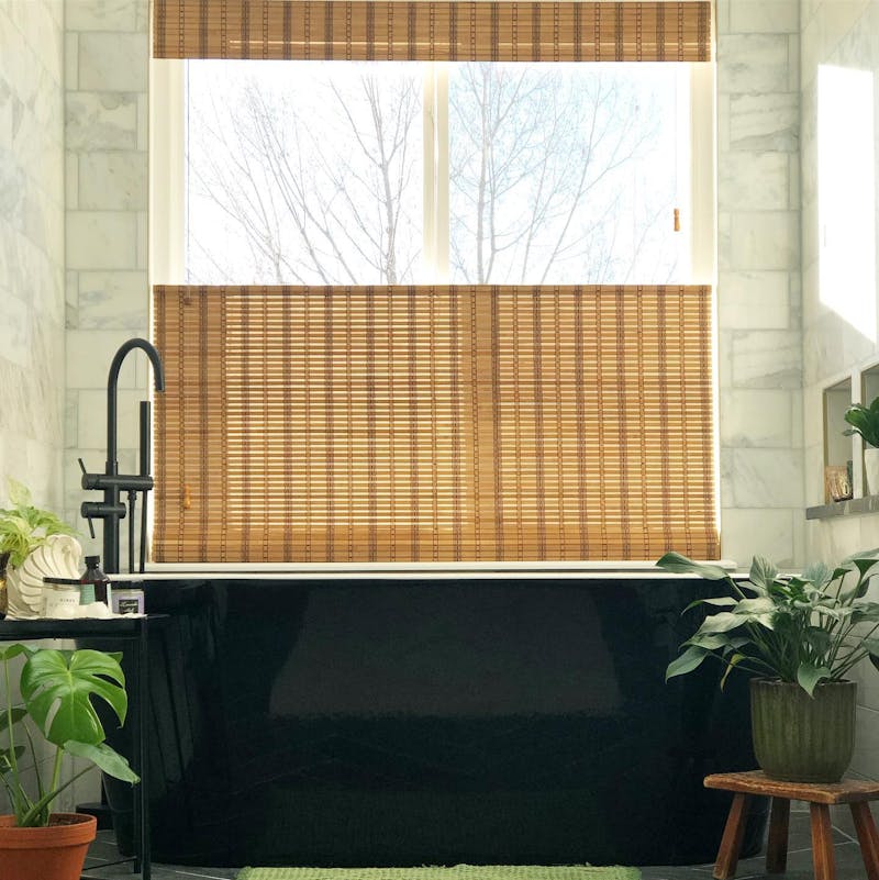 Large, black, free standing tub next to a window with a woven wood shades and surrounded by plants.