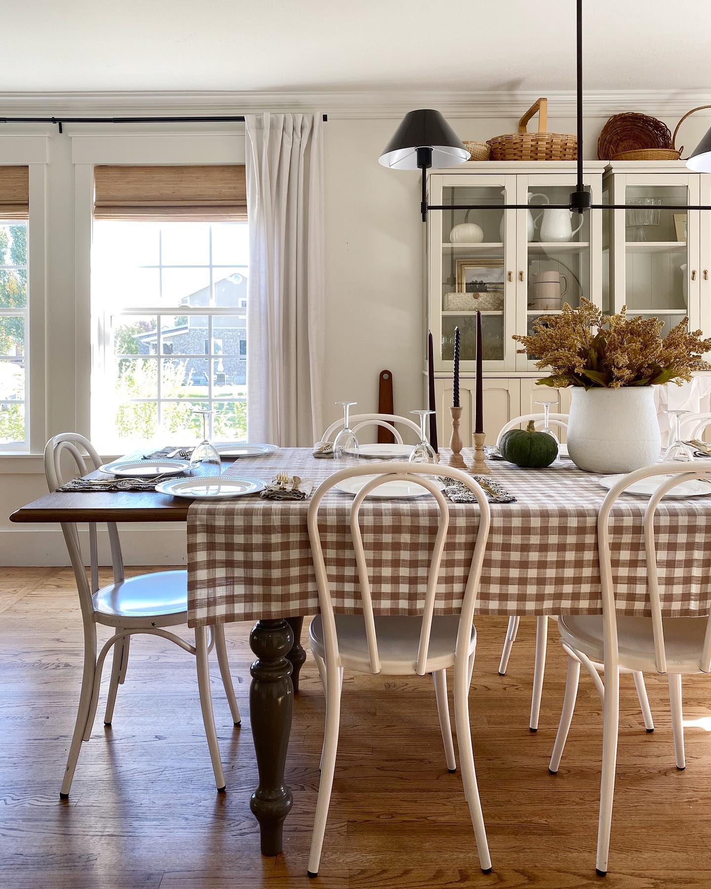 A dining table decorated for fall that is partially covered with a brown checkered tablecloth. There is fall home decor on the table. There are woven wood shade window coverings from Blinds.com on the windows. 