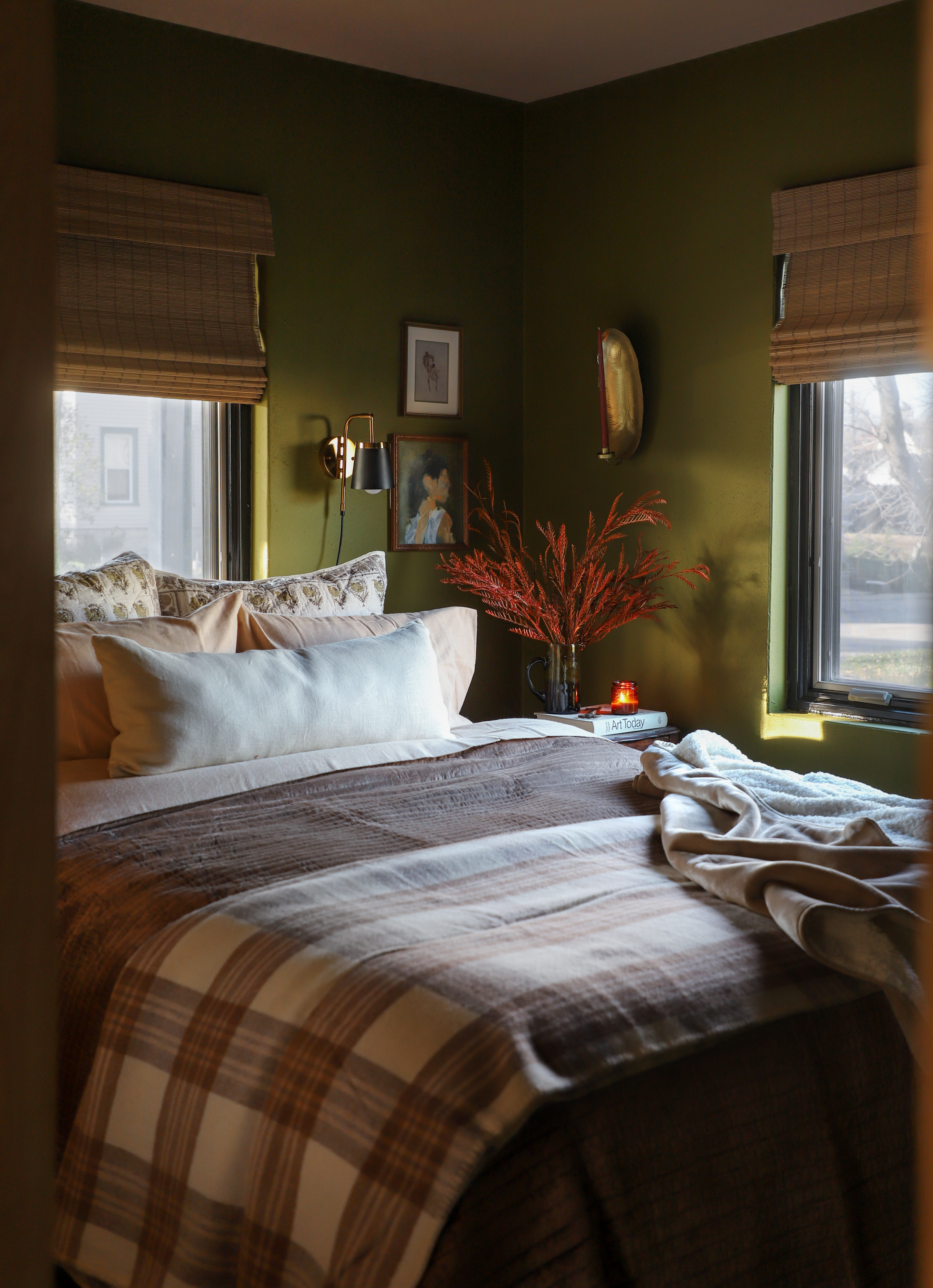 Fall bedroom decor. Bedroom with deep green walls and neutral bedding with a plaid pattern. Blinds.com woven wood shade window coverings are on the windows.