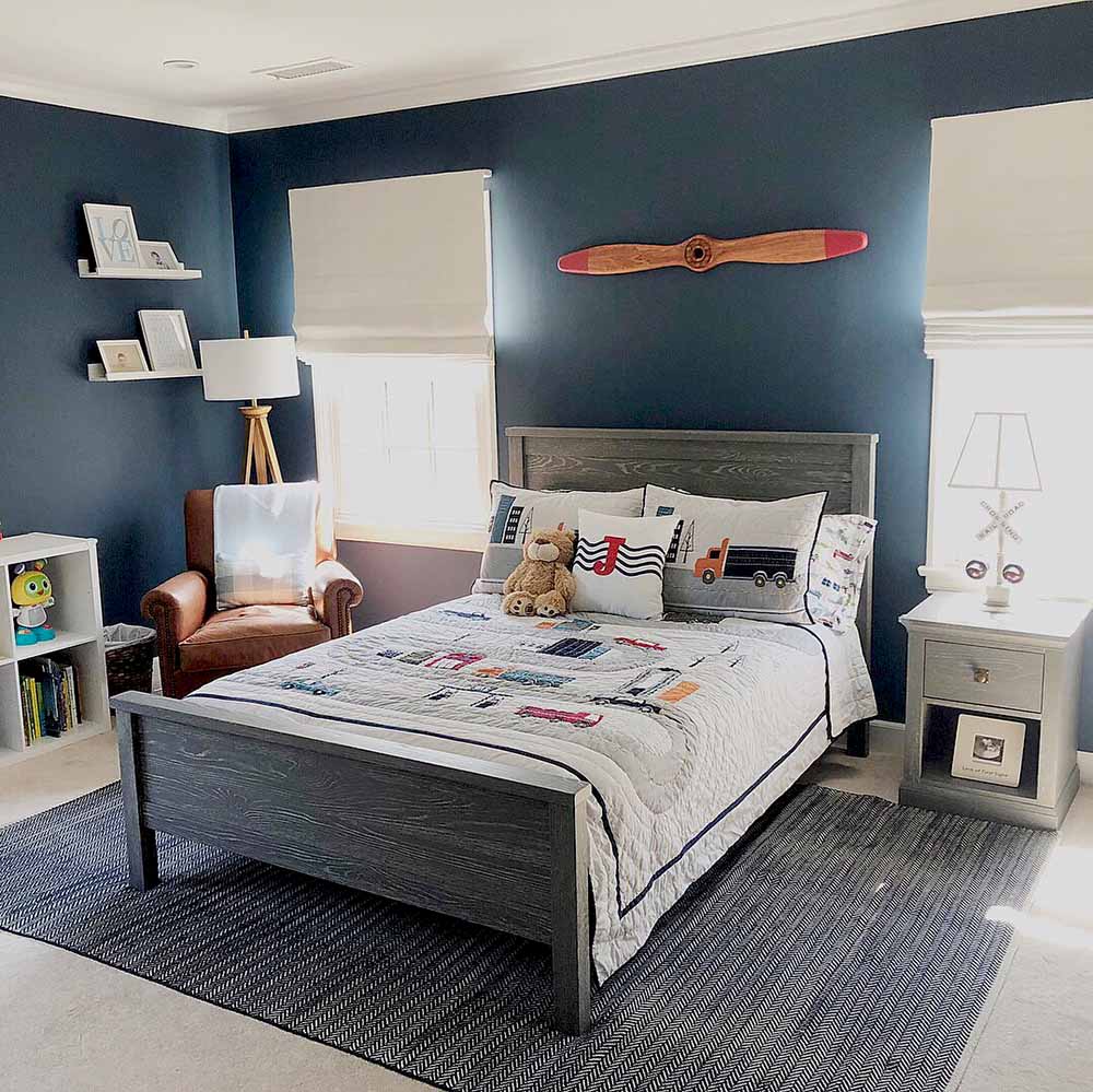 Young boy's bedroom with Classic Blue walls, quilted bedspread, two windows with beige roman shades and a decorative airplane propeller on the wall.