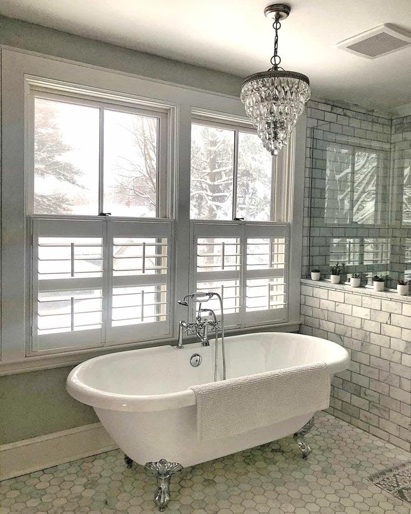 Contemporary tiled bathroom with shiny clawfoot tub and cafe height shutters over windows that look out onto a snowy field.