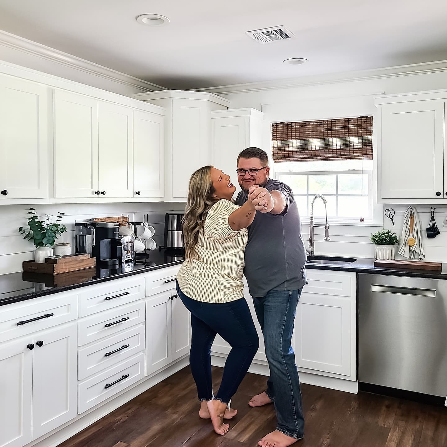 A couple dancing in the kitchen in front of a window with brown woven wood shades.