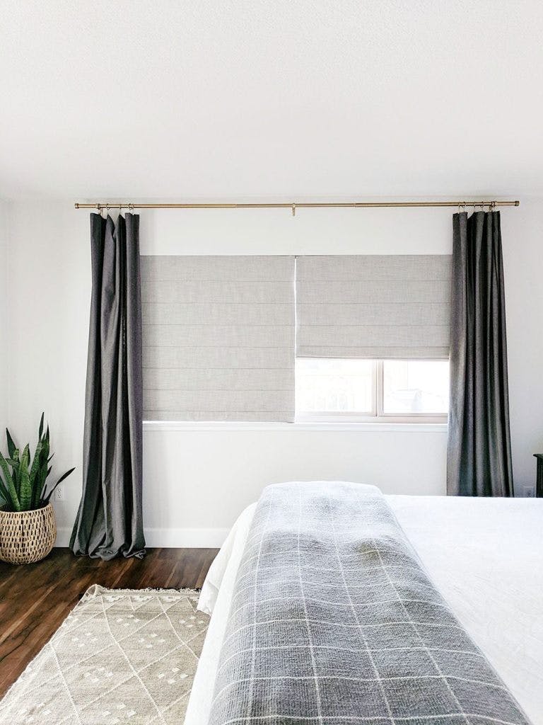 bedroom window with two beige roman shades and grey curtains with gold curtain rod