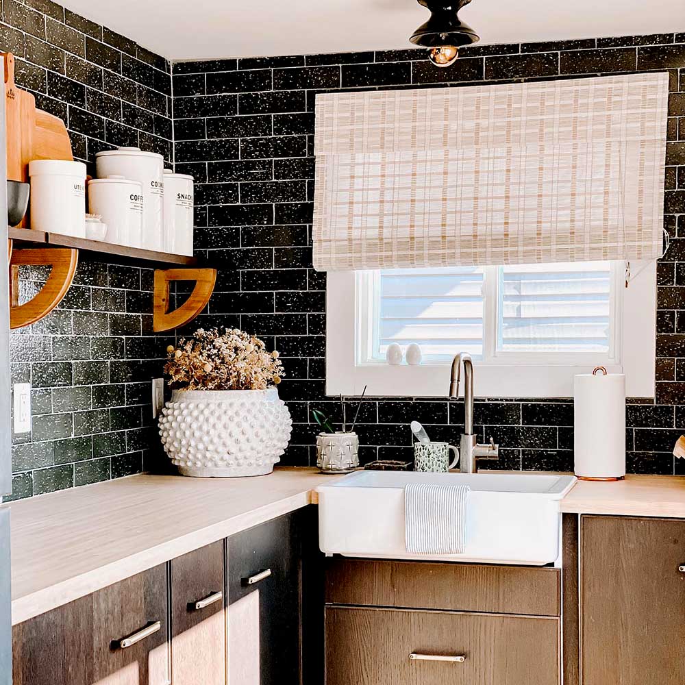Farmhouse kitchen sink with black subway tile backsplash, white countertops, wood shelving and beige roman shades on the window above the sink.